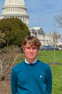 Conrad Gelber on the lawn in front of the U.S. Capitol, its dome rising behind him, wearing a teal sweater over a white collar.