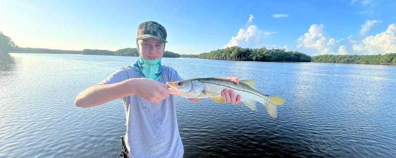 Conrad in a cap and sunglasses holding a snook across his chest on the deck of a boat, with calm water and a mangrove shoreline behind him under a clear sky.